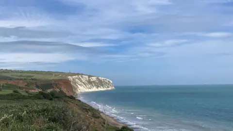 Ulani The white cliffs on the coast of the Isle of Wight in the distance. There are green fields on the left of the image and the sea on the right, with waves breaking at the shore below.