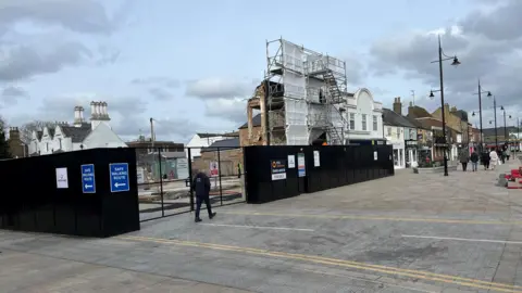 John Devine/BBC One section of a building is clad in scaffolding and white sheeting about 30 feet (9m) high. A black fence surrounds the partly demolished structure, some people are walking along the wide pavements, with ornate black lampposts visible too.