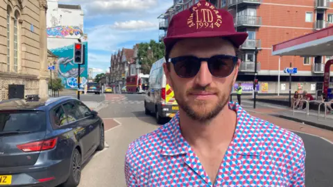 Hayden wearing a pink and blue patterned shirt, a burgundy cap and sunglasses. He is standing outside a petrol station inside the cordon. 
