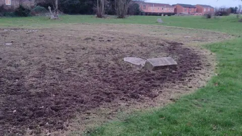 Durham Neighbourhood Wardens A large ring of trampled mud with a fallen headstone in the mud. Other headstones can be seen in the field further away.