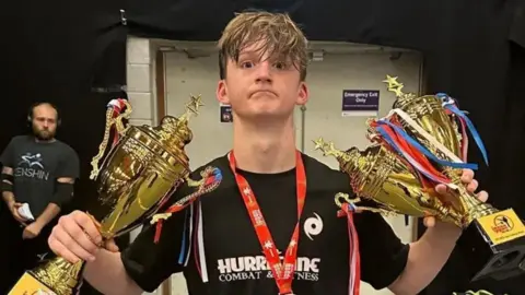 A teenage boy stands wearing a black t-shirt and holding three gold trophies and with a medal around his neck.