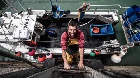 Buck Bennett Buck Bennett is smiling and climbing up a ladder on the side of a harbour wall from his boat which can be seen below. There is a fellow fisherman on the boat along with empty fishing boxes.