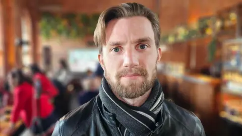 Hayden Bloomfield from 782 Marketing company pictured at the Copper Rivet Distillery in Chatham, Kent. Hayden has short hair, a short beard and is wearing a black scarf and black leather jacket. 