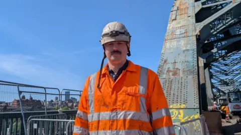 Taylor stands clad in bright orange high vis clothing in front of the rusted arch of the Tyne Bridge. The Newcastle skyline can be seen against the bright clear sky. 