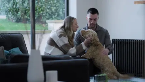 Stewarts Solicitors A woman with blonde hair and brown and white stripy jumper, pets a brown dog which has its paws on her lap. A man with dark hair and brown jumper is next to them

