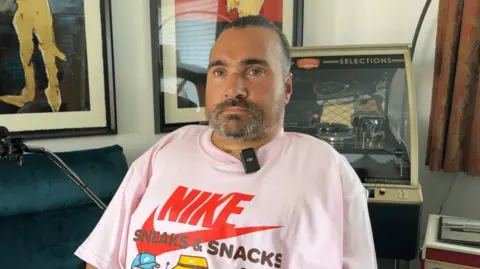 BBC A bearded man in a pink 'Nike Sneaks & Snacks' t-shirt sits in front of a green couch. Behind them are two framed artworks on the wall and a jukebox to the right.