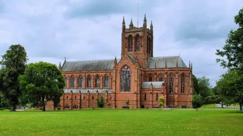 David Dixon The stone structured cathedral style Crichton Memorial Church, featuring a protruding tower and multiple windows, surrounded by gardens and trees on a cloudy day