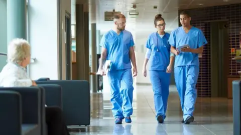 Getty Images A stock image of three healthcare professionals walking through what appears to be a hospital corridor, dressed in blue scrubs. An elderly patient looks on from a seating area in the foreground.