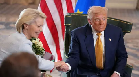 Getty Images Ursula von der Leyen, left, with short blonde hair and wearing a pale blue suit, smiles as she shakes hands with Donald Trump, wearing a blue suit and gold tie with a US pin, as they sit on leather chairs with EU and US flags behind them