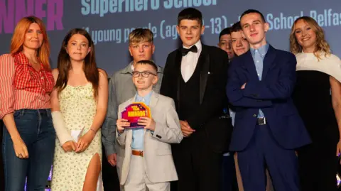 A group of nine people on a brightly lit stage. They are all dressed smartly and looking at the camera. The young boy in the middle of the group is wearing a grey suit and holding an award in his hands. 