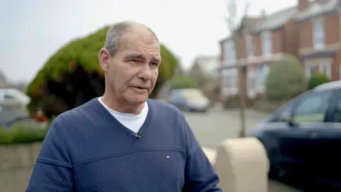 Steve, a man with greying hair cropped close, wearing a blue V-neck jumper over a white T-shirt, standing outside his home on the street where the attack occurred, with red-brick Victorian semi-detached houses behind him.