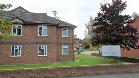 A two storey modern brick building with a small grassed area and low wall and a tree. There is a white sign that says Lotus Care: Cressington Court. 