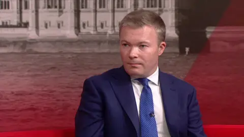 Bradley Thomas in the Politics Midlands studio in a blue suit and tie, with a white shirt. He is sitting on a red sofa