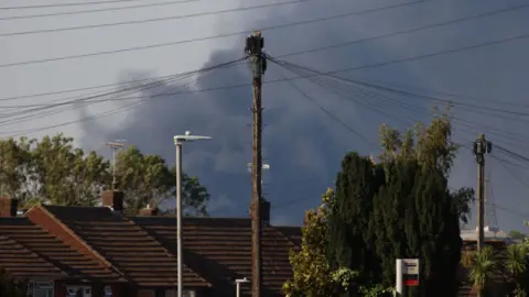 Stuart Woodward/BBC Clouds of black smoke rising into the sky above a foreground of houses and trees.