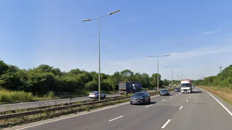 Google Drivers are coming towards the camera on a dual carriageway that has three lanes. On the other side of the road is another three lanes, with a lorry and car on the road. 