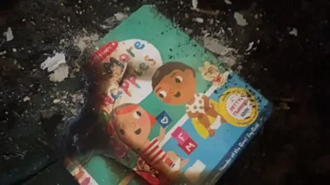 Little Wild Tribe The remains of a children's book with two cartoon children on the front, which has been badly burned.
