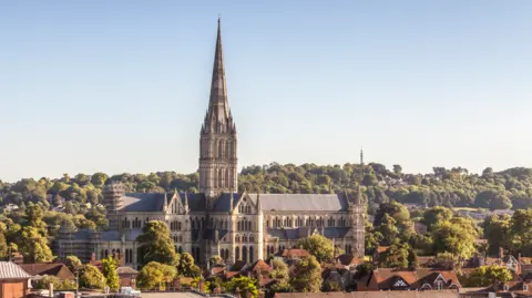 A wide shot of the Salisbury landscape including the cathedral with green trees surrounding it. There is a blue sky in the background.