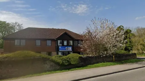 A Streetview image of Stoneyford Care Home, which is a two-storey brick building set behind a low wall with trees in the garden