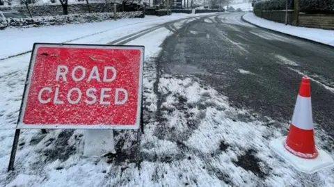Red 'Road Closed' on a road in Lancashire with snow and ice covering the road. A red and white traffic cone is to the right of the sign.
