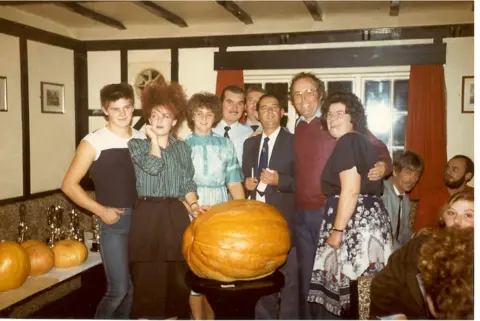 Kevin Fortey An old photo, shot and film, showing a group of men and women crowded in a dingey pub. They are surrounding a large pumpkin, placed on a wooden plinth.