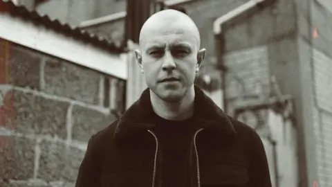 Skeet89 A black and white picture of Young Spencer. He has a shaved head and is wearing a dark jacketand top 