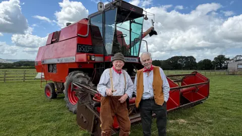 Pete Budd and Tommy Banner from the Wurzels stand in front of a combine harvester on a field.