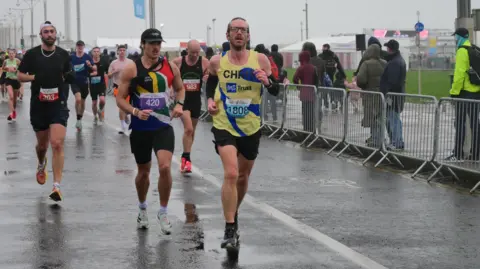 marathon-photos.com A group of runners is moving along a wet road during a race on a rainy day. Chris White is in the foreground, wearing a yellow and blue vest with the number 1808. He is running beside another runner in a black vest with a rainbow-coloured diagonal stripe and the number 420. Several other runners follow behind, while spectators and race marshals stand behind metal barriers on the right side of the road. The sky is grey and overcast, and the pavement is shiny from the rain.