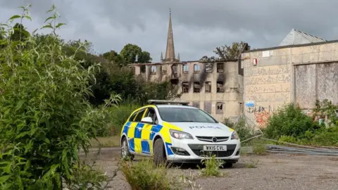 A police car in front of the Saxondale site which has large, black burn marks on the walls.