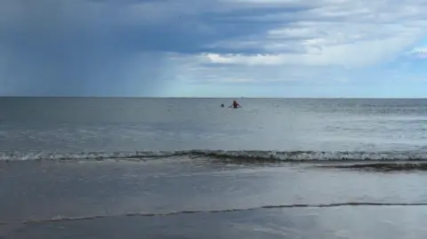 The silhouettes of two people swimming in the sea with waves lapping in the foreground and a moody skyline, a mix of blue sky, white clouds and a rain cloud