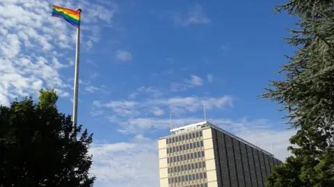 Norfolk County Council A rainbow flag flying from a flagpole. To the right of the image is a multi-storey rectangular building with multiple windows. 