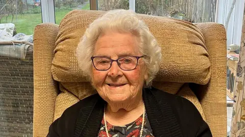 PA Media Olga Hopkins wearing a black cardigan over a flowery top. She has short grey hair and is smiling while sat on a cream armchair.