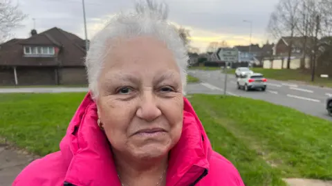 Ruth Grant who has short white hair and is wearing a bright pink jacket. She is stood on a pavement with grass, trees, a road with cars and houses in the background.