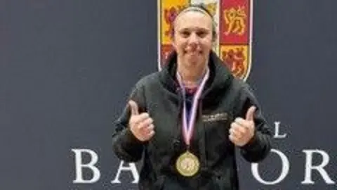 A fair-haired woman in a black hoodie wears a gold medal with her thumbs up.