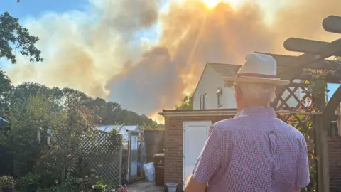 Robby West/BBC Elderly man wears a straw hat and stands with his back to the camera. He is in his garden and ahead of him are plants and a greenhouse. In the near distance, orange smoke blows in the wind. 