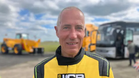 A man looks towards the camera wearing a yellow and black driving suit with the "JCB" logo on the front