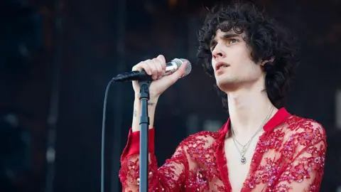 A singer with long dark hair, wearing a see-through red shirt, hold a microphone
