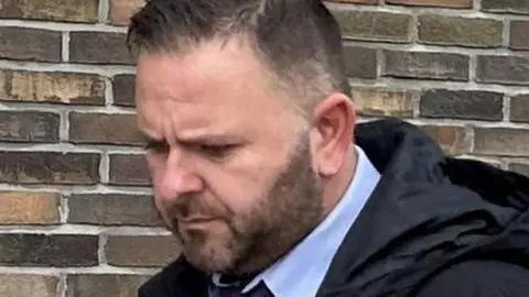 PC Paul Higgins, with short dark hair and beard, and looking down. He is wearing a suit and coat walking past a brick wall. 