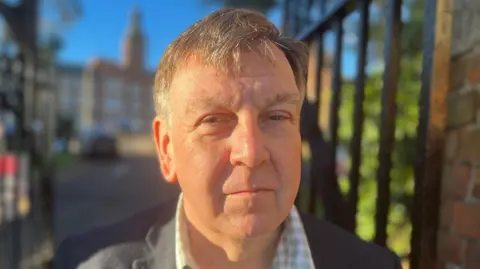 Sir John Whittingdale standing in front of iron gates, which are blurred. He has short grey hair and is wearing a chequered shirt.