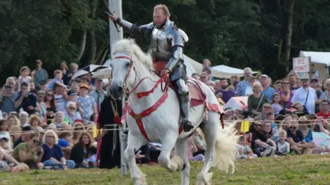 A man in armour holding a sword, riding a white horse while crowds watch
