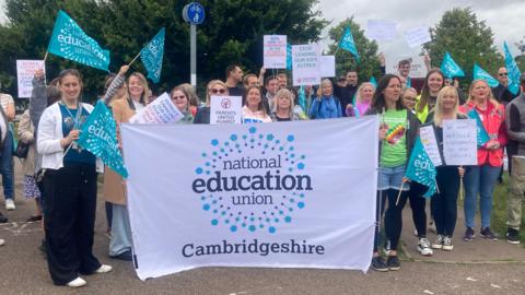 Picket line members waving flags and holding a banner which reads: "National Education Union Cambridgeshire"