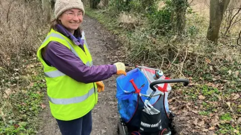 A woman wearing a high viz jacket is pictured using a litter picker to put waste inside a plastic rubbish bag.