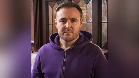 ITV/Coronation Street Tyrone Dobbs played by Alan Halsall wears a purple hoodie and is staring into the camera lens with a straight face.
