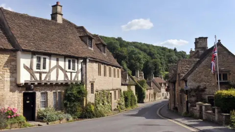 A wide shot of a street passing through Castle Combe in the Cotswolds. Timber-framed houses made of traditional stone are seen on either side of the road and there is a Union Jack flag on a pole in one of the gardens. Woodland is visible in the distance