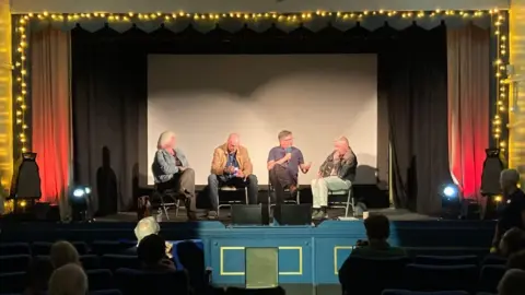 Lucem House Community Cinema Four people are sitting on a chairs on a stage with microphones while a number of people in the audience are watching