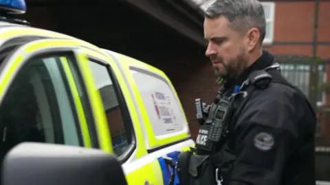 PC Dalby opens door ofyellow and blue liveried police car