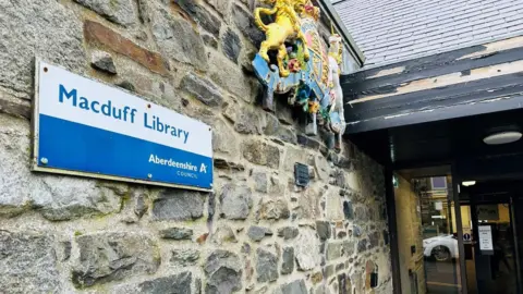 Building with Macduff Library / Aberdeenshire Council sign in blue and white on the wall, with a bright crest higher up the wall, and library doors.