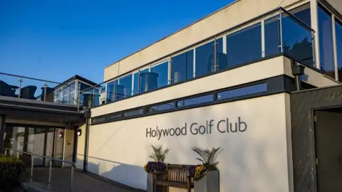 PA Media The front of Holywood Golf Club, a beige stone building, with dark lettering saying the words Holywood Golf Club on the front. It has glass boxes on top of the building and there is a brown bench at the front.