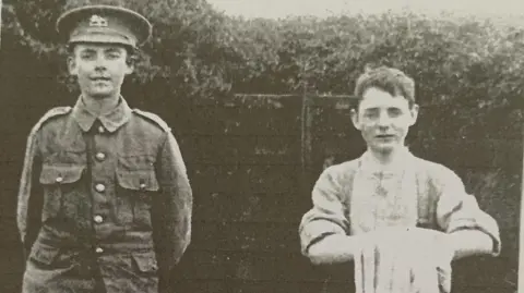 A black and white image of Edward and Joseph Johnston. Edward is wearing a military uniform and hat and standing with his hands behind his back. Joseph stands next to him. He wears a shirt and holds an item of clothing folded over his hands. Behind them is a wooden garden fence with a hedge poking over the top.