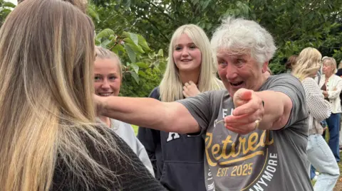 An woman with short white hair wears a T-shirt which says "retired" while offering a hug to a young woman who is standing in a group of teenagers.