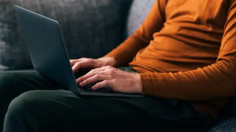 Getty Images A stock image showing a man from the shoulders down sitting on a grey sofa with a laptop on his lap. He is wearing an orange top and a green pair of trousers.